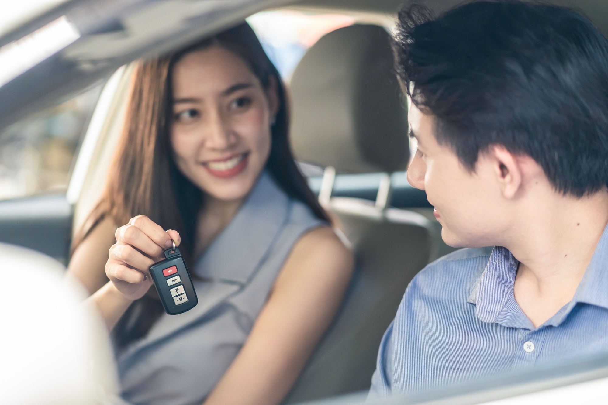 Asian woman showing car remote key to man friend sitting in vehicle at car rental company together